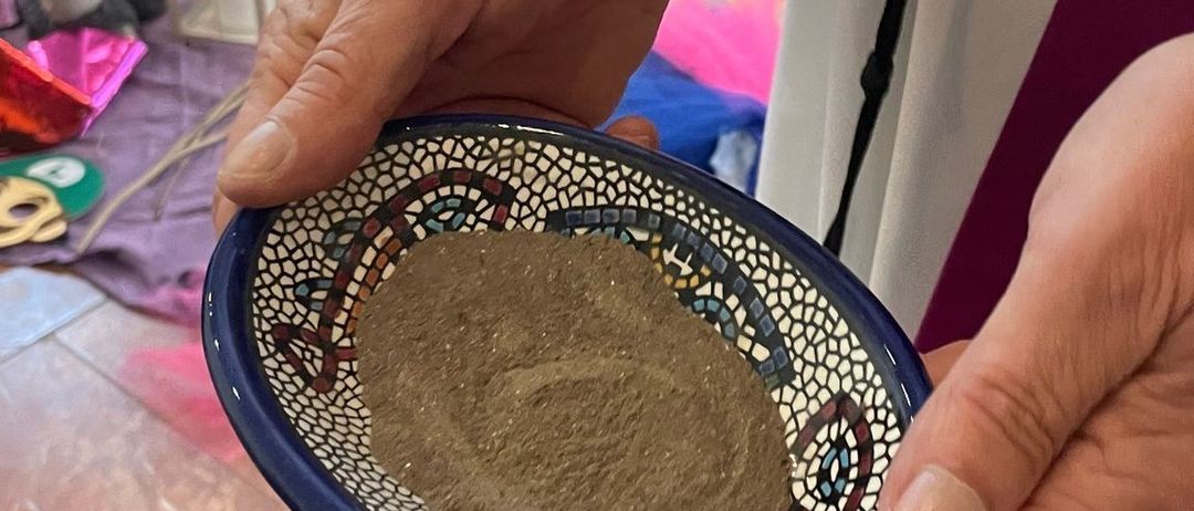 Eine Person hält eine Schüssel mit braunem Pulver, mit einem gemusterten Rand, in den Händen. Der Hintergrund zeigt eine brennende Kerze, einen Laternen und einen gefliesten Boden.