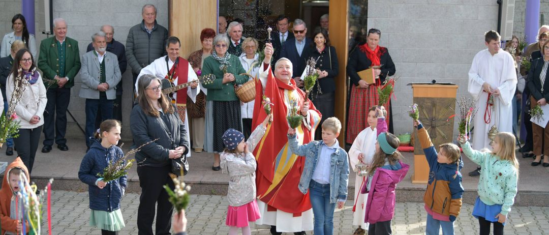Eine religiöse Zeremonie im Freien mit einem Priester, der Blumen hält, umgeben von Kindern und Erwachsenen, einige halten Blumen und andere beobachten.