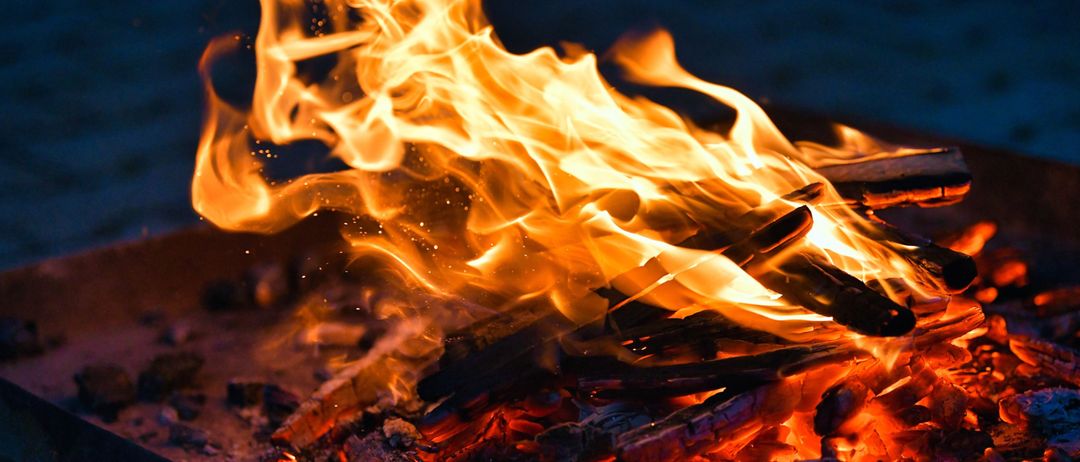 Ein Feuer brennt hell, die Flammen tanzen in der Nacht, umgeben von verkohltem Holz und glühenden Glut.