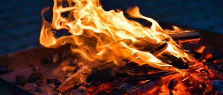 Ein Feuer brennt hell, die Flammen tanzen in der Nacht, umgeben von verkohltem Holz und glühenden Glut.