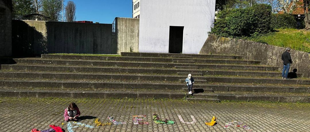 Zwei Kinder spielen auf den Stufen eines weißen Gebäudes. Eines ordnet bunte Buchstaben auf dem Boden an. Ein kleiner Baum und ein Laternenpfosten befinden sich auf der rechten Seite.