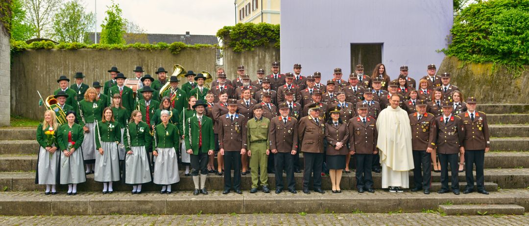 Eine Gruppe uniformierter Personen, darunter Militärangehörige und Musiker, steht in Formation auf einem Betonpodest. Dahinter sind Gebäude und Grünflächen zu sehen.