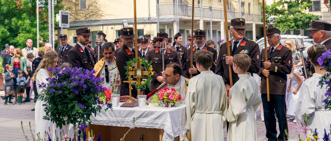 Eine feierliche Zeremonie mit einem Priester am Altar, umgeben von Militäroffizieren und zwei jungen Akolythen. Der Altar ist mit Blumen und Kerzen geschmückt, vor dem Hintergrund von Gebäuden.