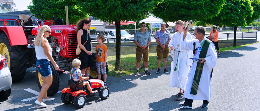 Ein Junge fährt einen roten Spielzeugtraktor, während ein Priester und sein Ministrant auf der Straße stehen, während Erwachsene und Kinder sich versammeln.