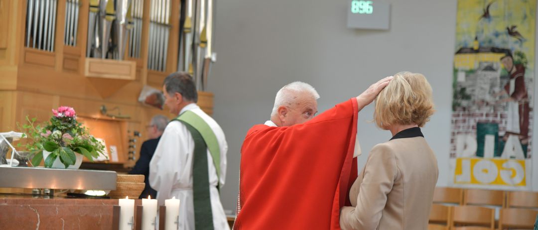 Ein Priester in einem roten Gewand legt während eines Gottesdienstes seine Hände auf den Kopf einer Frau in einem beigen Blazer. Kerzen sind angezündet, und eine digitale Uhr zeigt 896 an der Wand.