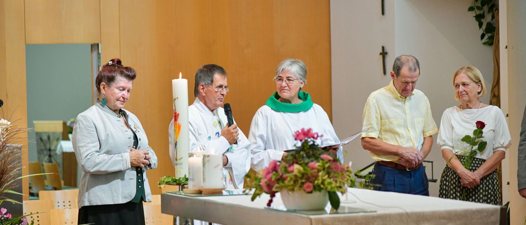 Vier Personen, darunter eine Frau und drei Männer, stehen vor einem Altar. Sie tragen weiße Gewänder und halten Kerzen. Ein Mikrofon steht vor einem Mann.