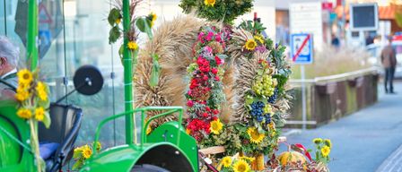 Ein grüner Traktor, geschmückt mit Blumen- und Erntedekorationen, darunter Kürbisse, Mais und Trauben, auf der Straße geparkt.