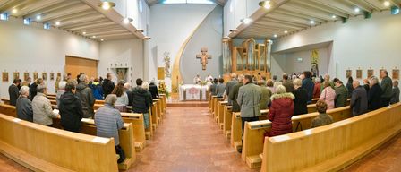 Eine Gruppe von Menschen versammelt sich in einer Kirche und schaut auf den Altar, wo ein Priester eine Zeremonie durchführt. Die Kirche ist gut beleuchtet mit hölzernen Kirchenbänken und einem Kreuz an der Wand.