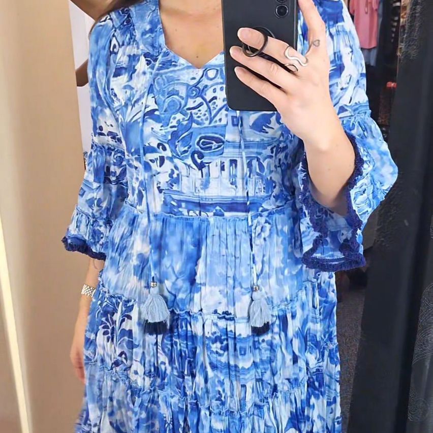 Eine Frau macht ein Selfie in einem blauen Kleid mit komplizierten Designs, hält ein Telefon und trägt eine silberne Uhr.
