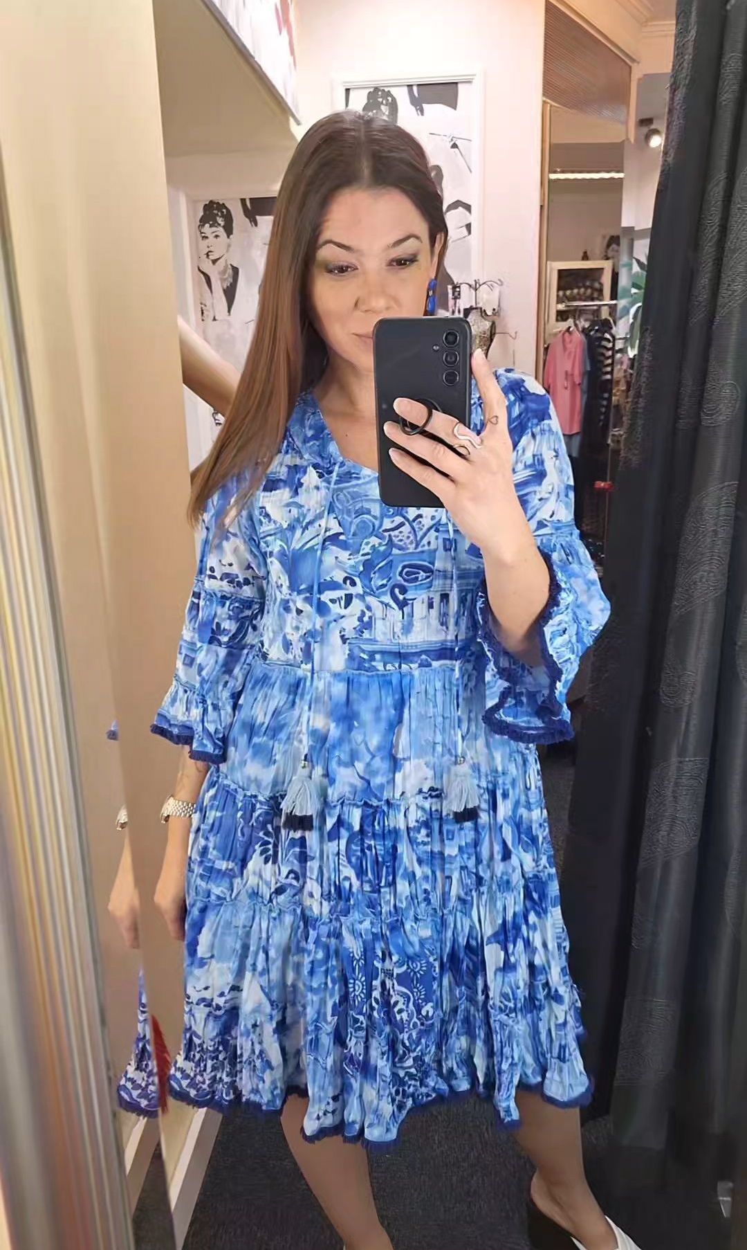 Eine Frau in einem blauen Kleid mit Blumenmuster macht ein Selfie vor einem Spiegel. Hinter ihr hängen Kleider an einem Bügel.
