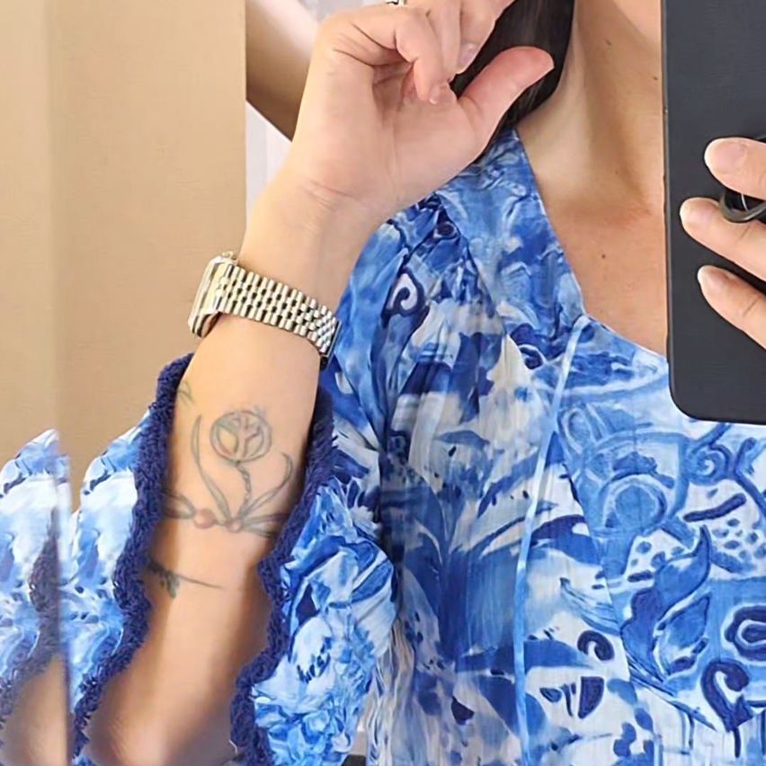 Eine Frau hält ein Telefon und untersucht ein Tattoo an ihrem Arm. Das Tattoo ist eine Blume mit vielen Details. Sie trägt ein blau-weißes Blumenkleid.