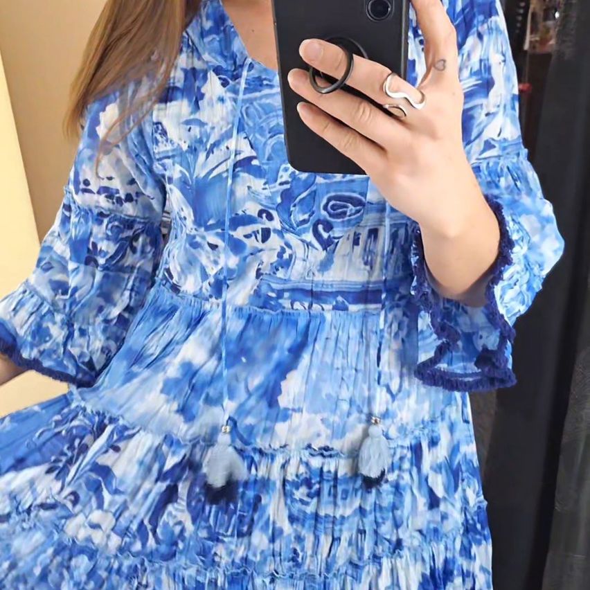 Eine Frau macht ein Selfie in einem blauen Blumenkleid, hält ein schwarzes Smartphone mit einem Ring und einem Armband am Handgelenk.