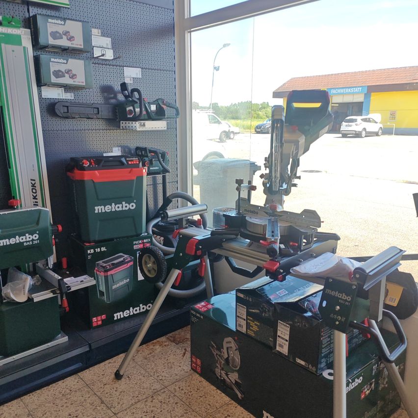 Ein Schaufenster mit verschiedenen Metabo-Werkzeugen. Mehrere Werkzeuge und Akkus sind an den Regalen und am Boden ausgestellt. Ein geparktes weißes Auto ist durch das Glasfenster sichtbar.