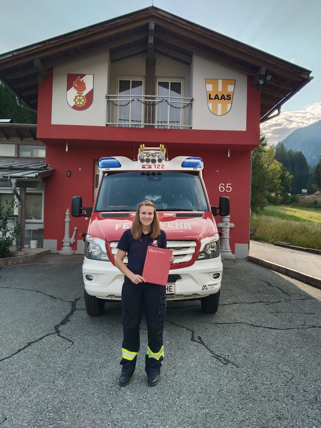 A woman in a uniform stands in front of a red fire truck with the number 122 and the word 'Feuerwehr' on the front. Behind her is a red building with a balcony and a logo on the wall.