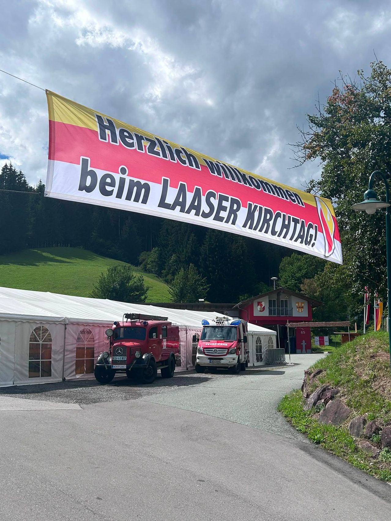Ein großes Banner mit der Aufschrift 'Herzlich willkommen beim LAASER KIRCHTAG!' hängt über zwei Feuerwehrwagen, die vor einem roten Gebäude mit einem weißen Zelt in der Nähe geparkt sind.