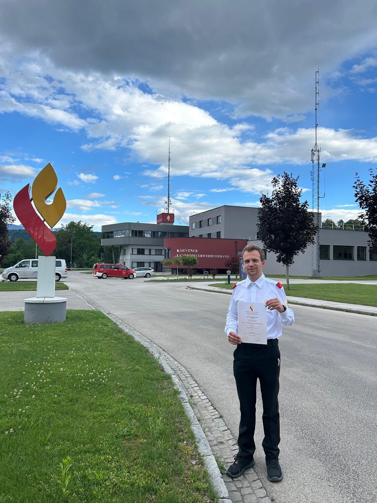 A man in uniform stands in front of a building with a sculpture. A red van and a white van are parked nearby. The sky is blue with some clouds.