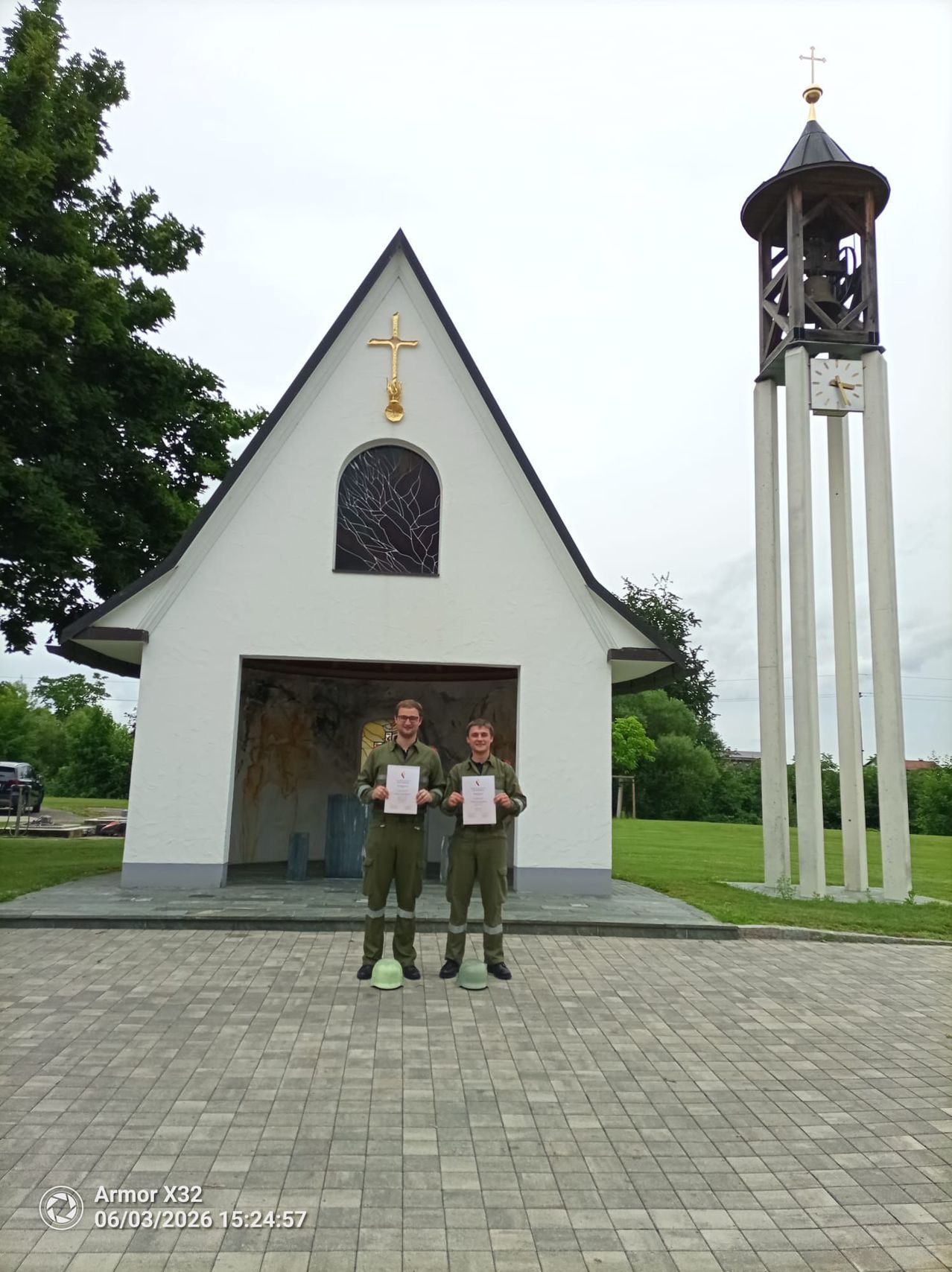 Two men in green uniforms stand in front of a small church with a bell tower. They hold certificates and smile for the camera.
