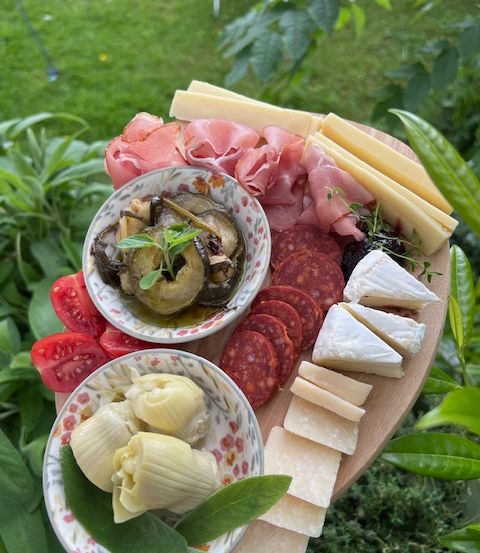Ein Holzbrett mit verschiedenen Lebensmitteln, darunter geschnittenes Fleisch, Käse, Artischocken und Auberginen, vor einem Hintergrund von Grün.