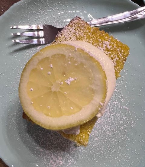 Eine Zitronenscheibe auf einem Kuchen, bestreut mit Puderzucker, liegt auf einem Teller mit einer Gabel.