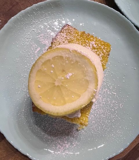 Eine Zitronenscheibe liegt auf einem quadratischen Stück Kuchen auf einem weißen Teller, bestreut mit Puderzucker. Der Hintergrund ist ein Holztisch.