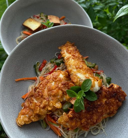Zwei Schüsseln mit knusprigen Fischfilets auf Nudeln mit Gemüse und einer Basilikumblattgarnitur, eine im Vordergrund und eine im Hintergrund.