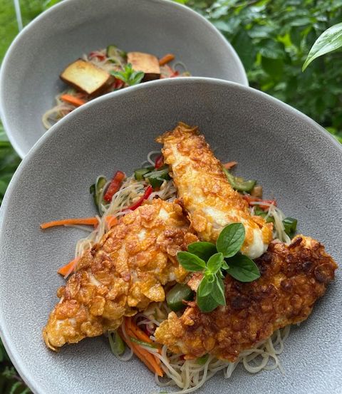 Zwei Schalen mit knusprigen Fischfilets und Gemüse auf Nudeln mit Basilikumblättern, serviert auf einem Holztisch mit Pflanzen im Hintergrund.