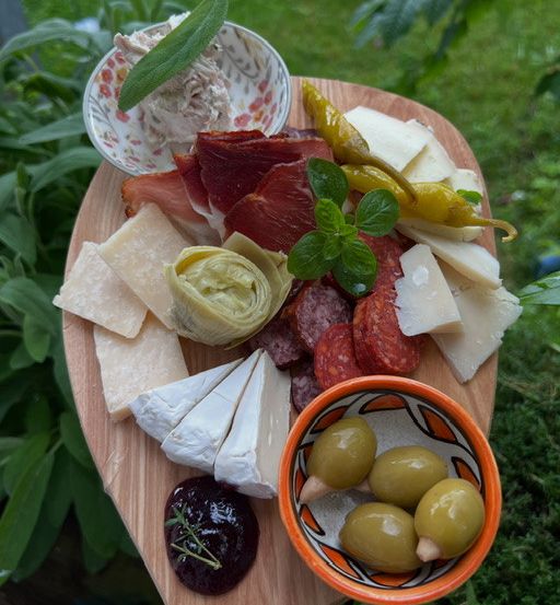 Ein Holzbrett zeigt eine Auswahl an gereiften Fleischsorten, Käse, Oliven und Kräutern. Ein kleines Schüsselchen mit grünen Oliven steht rechts.