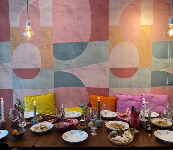 Ein Esstisch ist mit Tellern, Schalen und Weingläsern gedeckt. Bunte Kissen und Kerzen sind an der Wand.