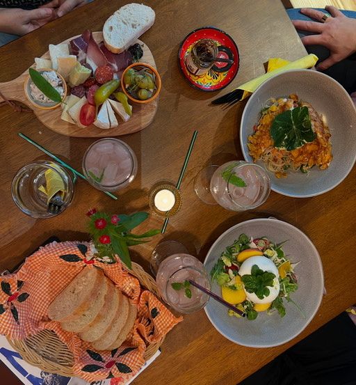 Ein Holztisch mit verschiedenen Gerichten, Getränken und Utensilien. Teller mit Salaten, ein Käsebrett und Oliven sind vorhanden. Gläser enthalten Getränke und eine Kerze ist angezündet.