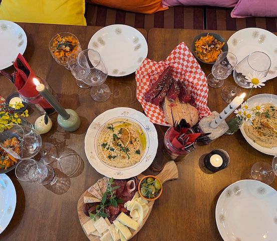 Ein Holztisch ist mit Tellern, Schüsseln, Weingläsern und Kerzen gedeckt. Es gibt verschiedene Gerichte auf dem Tisch, darunter Hummus, Käse und Fleisch.