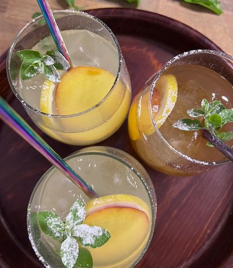 Drei Gläser mit Pfirsich-Cocktails und bunten Strohhalmen, garniert mit Minzblättern und Pfirsichscheiben, stehen auf einem Holztablett.