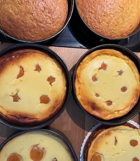 Fünf gebackene Waren in schwarzen Formen: zwei ganze Kuchen und drei Käsekuchen mit Löchern und Brandflecken. Jedes Element steht auf einer Holzoberfläche.