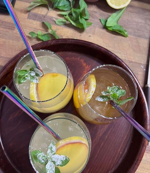 Drei Gläser eines Pfirsich-Minz-Cocktails stehen auf einem Holztablett, garniert mit Pfirsichscheiben und Minzblättern, mit einer Zitronenscheibe an der Seite.