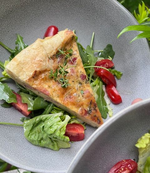 Ein Stück Quiche mit Schinken und Käse wird auf einem Teller serviert, begleitet von einem Salat aus grünen Blättern und roten Tomaten.