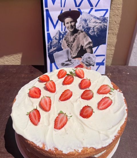 Ein mehrschichtiger Kuchen mit weißer Glasur und Erdbeeren steht auf einem Tisch. Dahinter befindet sich ein Poster mit einer Frau und Bergen.