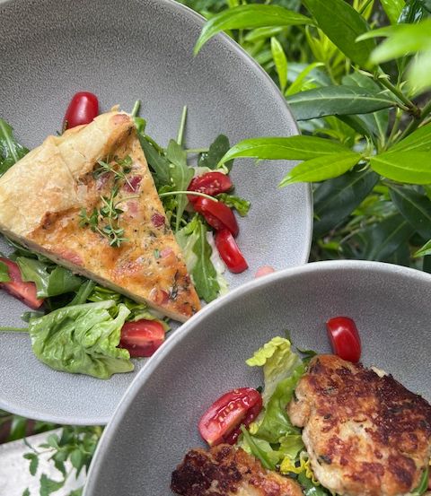 Zwei Teller mit Essen, einer mit einem Stück Quiche und Salat, der andere mit einem Hähnchenschnitzel und Salat, stehen auf einem Tisch draußen vor grünem Hintergrund.