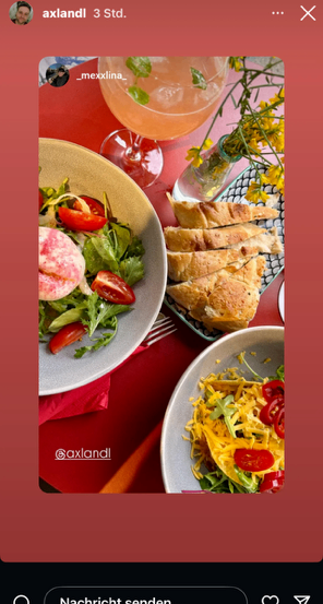 Ein Foto zeigt einen Tisch mit drei Gerichten: ein Salat, Brot und ein Gericht mit Käse und Tomaten. Dazu ein Glas rosa Getränk mit Minzblättern und ein Blumenarrangement.