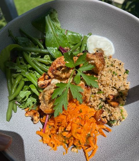 Ein Teller mit gegrilltem Fleisch, grünen Bohnen, Salat, Karotten und Couscous, garniert mit Petersilie, aufgenommen im Freien.