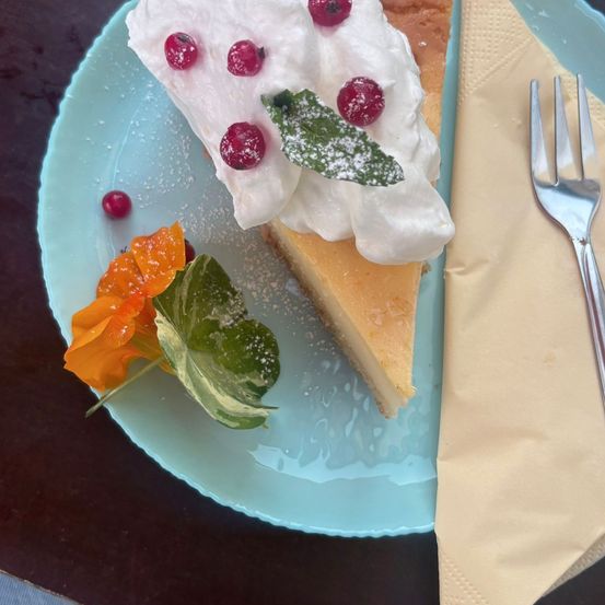 Ein Stück Käsekuchen auf einem hellblauen Teller. Es ist mit Schlagsahne garniert, mit roten Beeren verziert und mit einer orangefarbenen Blume und grünen Blättern verziert. Eine Gabel ruht an der Seite des Tellers.