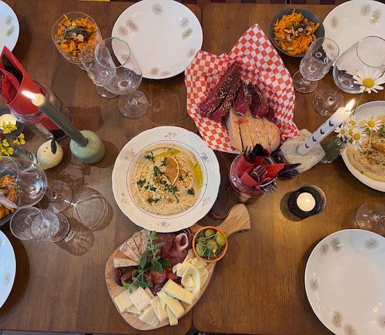 Ein hölzerner Tisch mit mehreren Gerichten und Getränken, darunter Hummus, Fleisch, Käse und Weingläser. Teller, Schüsseln und Servietten sind angeordnet. Kerzen sorgen für eine warme Atmosphäre.
