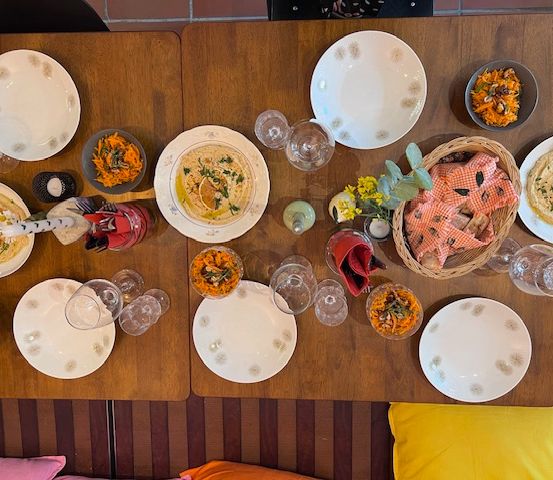 Ein Holztisch ist mit verschiedenen Gerichten wie Hummus und Salaten dekoriert, umgeben von Tellern und Gläsern, mit dekorativen Elementen wie Blumen und Servietten.