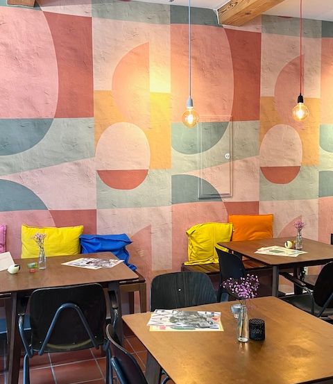 Das Innere eines Restaurants mit farbenfrohen abstrakten Wandkunst. Holztische und Stühle sind angeordnet, mit verschiedenen Kissen und einer Vase mit Blumen.