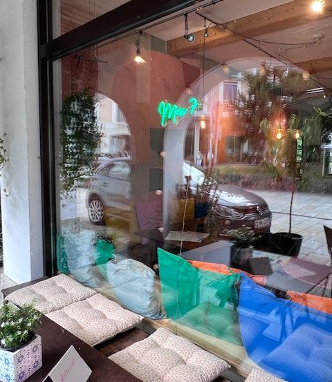 Ein modernes Schaufenster mit Neon-Leuchtreklamen und farbenfrohen Kissen. Autos sind draußen geparkt, und es gibt einen Topf mit Pflanzen und einen Tisch mit einer Karte darauf.