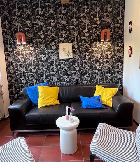 Ein Wohnzimmer mit einem schwarzen Sofa, das mit bunten Kissen geschmückt ist. Ein weißer Tisch mit einem Blumenvase steht zwischen zwei gestreiften Stühlen. Die Wände sind mit schwarzem Tapetentapeten mit Palmen und Tieren bedeckt.