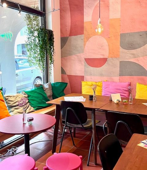 Ein gemütliches Café mit farbenfrohen Tischen und Stühlen. Die Wände sind in abstrakten Formen bemalt. Die Bank ist mit leuchtenden Kissen geschmückt. Ein Auto ist draußen geparkt.