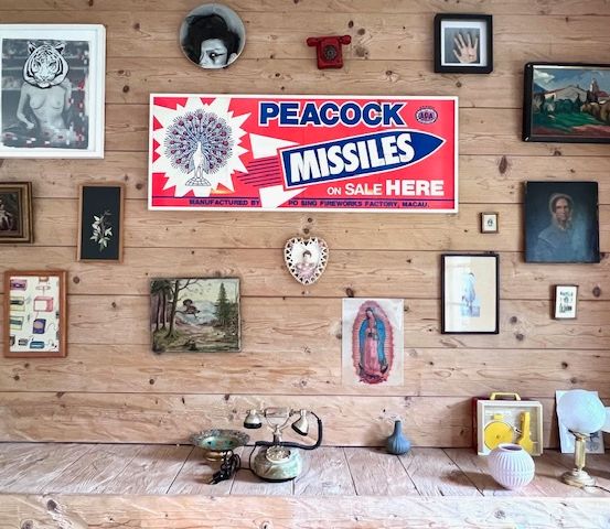 An einer Holzwand hängen verschiedene gerahmte Bilder und Poster, darunter ein Schild für Peacock Missiles zum Verkauf. Ein Telefon, eine Vase und andere Gegenstände stehen auf einem Holzregal.