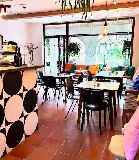 Ein Innencafe mit gefliestem Boden und Stühlen um Tische herum, einer Theke mit Flaschen und großen Fenstern mit Pflanzen.