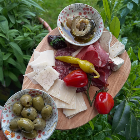 Ein Holzbrett zeigt verschiedene Käsesorten, Schinken, Oliven und Tomaten, mit einer Schüssel marinierter Zwiebeln in der Ecke.