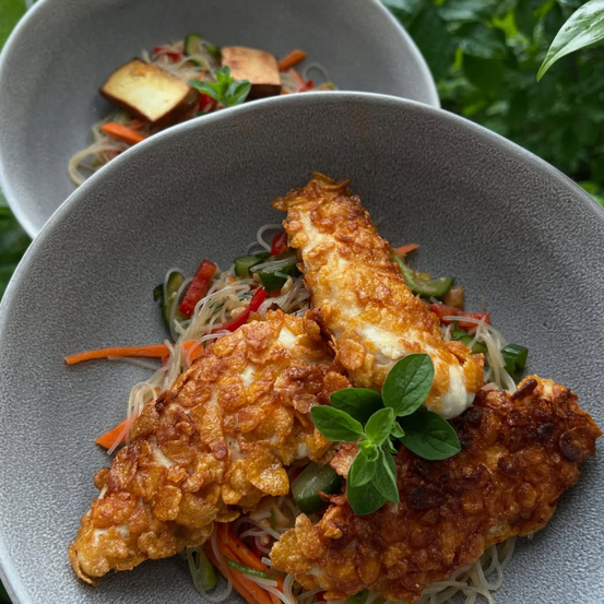 Ein Teller mit gebratenen Fischfilets, Gemüse und Nudeln. Der Fisch ist knusprig und goldbraun. Das Gericht hat eine grüne Kräutergarnitur.