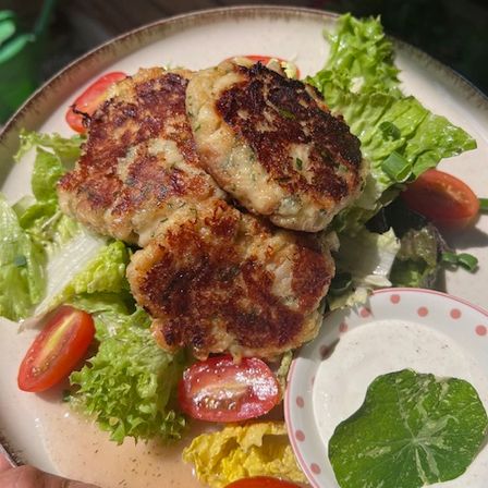 Ein Teller mit drei gebratenen Patties auf einem Bett aus Salat, begleitet von einer kleinen Schüssel grüner Sauce und geschnittenen Tomaten.